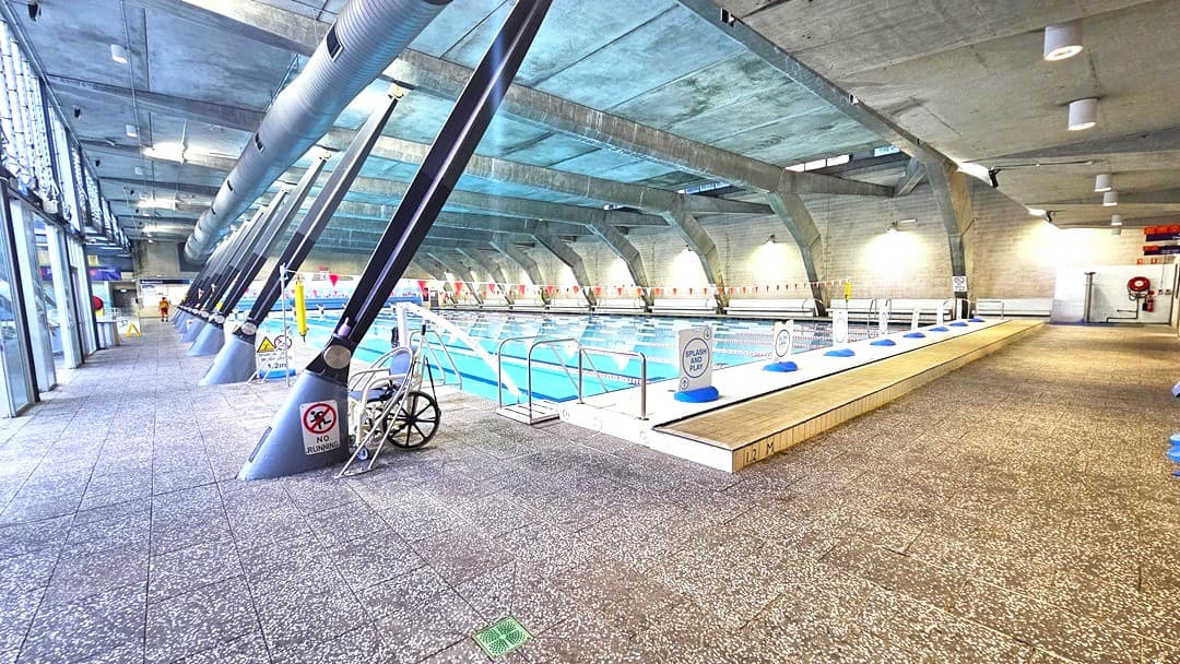 Indoor swimming pool with a tiled floor and a concrete ceiling. On the left, large windows allow natural light in, and there are several angled support beams. A wheelchair is parked near the poolside. The pool is in the centre, with lane dividers and safety signs along the edge. On the right, a walkway runs parallel to the pool, with benches and a lifebuoy on the wall. The space is well-lit with overhead lights.