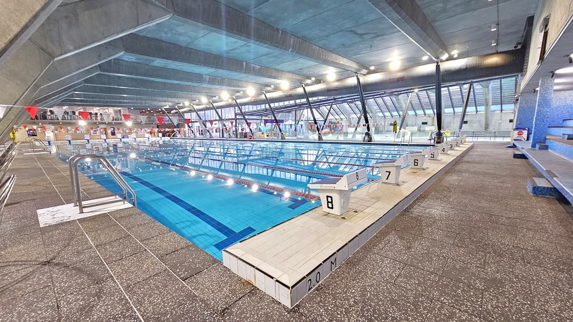An indoor swimming pool with multiple lanes, each marked with starting blocks numbered 1 to 8. The pool is surrounded by a textured, non-slip deck. On the left, there are metal handrails leading into the water. The ceiling is high with exposed beams and bright lighting. Large windows on the right allow natural light to enter. Benches are visible on the right side, providing seating for spectators. The pool area is spacious and well-lit.
