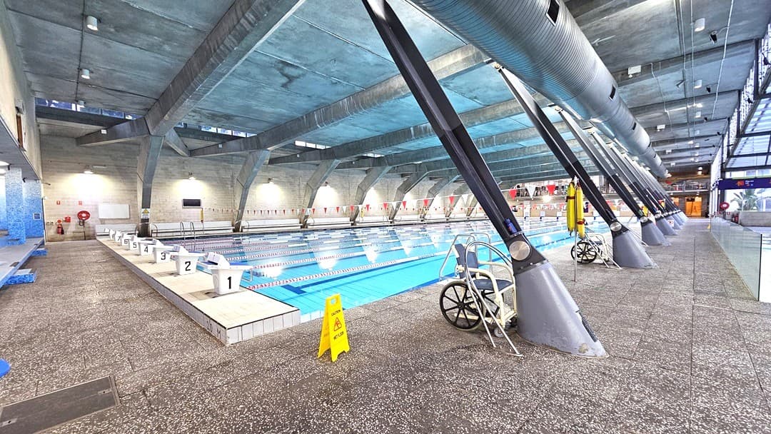 Indoor swimming pool with multiple lanes, starting blocks numbered 1 to 6 on the left. The pool is surrounded by a textured, non-slip floor. On the right, a wheelchair and a yellow caution sign are visible. Large, angled metal beams support the ceiling, which is high and industrial in style. The background features a wall with lights and a lifebuoy. The far end of the pool area has glass panels and a staircase leading to an upper level.