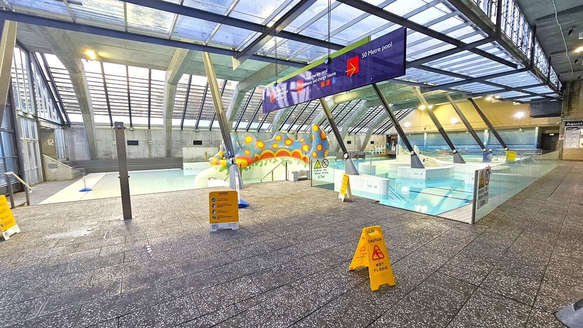 Indoor swimming pool area with a glass ceiling and large windows. In the centre, a colourful, caterpillar-shaped water feature is visible. The pool is divided into sections, with a sign above indicating a 50-metre pool. Yellow caution signs are placed on the wet tiled floor in the foreground. The area is well-lit with overhead lights, and there are metal railings and steps leading into the pool on the left.