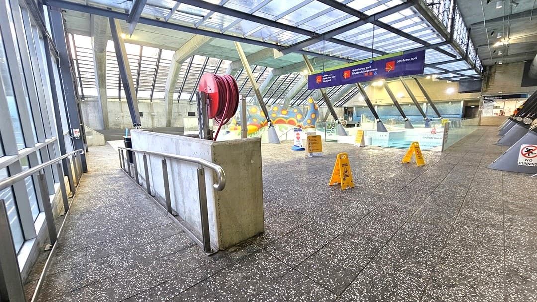 A spacious indoor area with a sloped walkway on the left, featuring metal railings. The floor is speckled grey, and the ceiling is made of glass panels supported by metal beams. In the centre, a red hose reel is mounted on a low wall. Brightly coloured caution signs are scattered around, and a large blue sign hangs from the ceiling. The background has a colourful mural and more glass walls, with a "No Smoking" sign visible on the right.