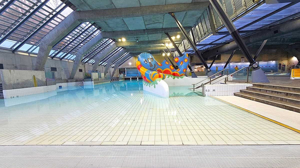 An indoor swimming pool with a colourful, abstract sculpture resembling an octopus in the centre. The pool has a shallow tiled area in the foreground, leading into deeper water. On the right, there are steps with handrails leading into the pool. The ceiling features large, slanted glass panels supported by metal beams, allowing natural light to enter. The walls are concrete, and there are additional seating areas and signage visible in the background.