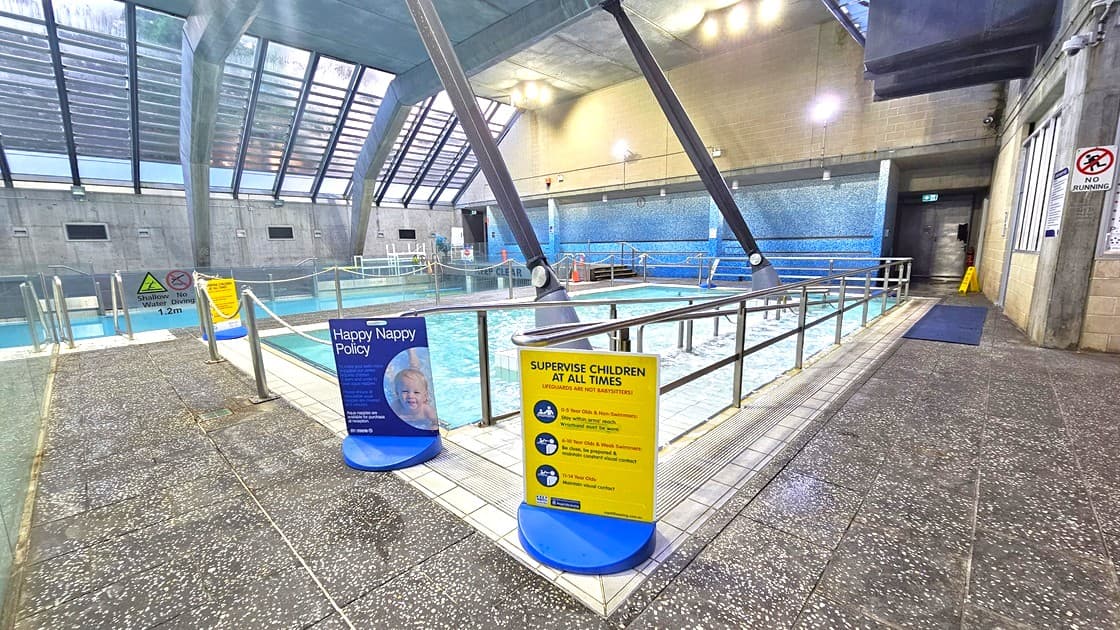 Indoor swimming pool area with a tiled floor and a high, slanted glass ceiling. In the foreground, there are signs about pool policies, including a "Happy Nappy Policy" and a reminder to "Supervise Children at All Times." The pool is surrounded by metal railings, and there are large structural beams supporting the ceiling. The walls are a mix of concrete and blue tiles. A corridor is visible on the right, leading to other areas of the facility.