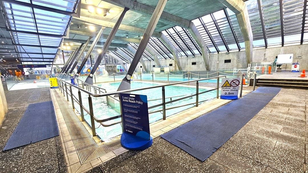 Indoor swimming pool area with a sloped glass ceiling supported by metal beams. The pool is surrounded by metal railings and has signs indicating safety policies and depth. Blue non-slip mats are placed on the tiled floor around the pool. In the background, there are orange cones and additional pool equipment. The lighting is bright, with multiple ceiling lights illuminating the space.