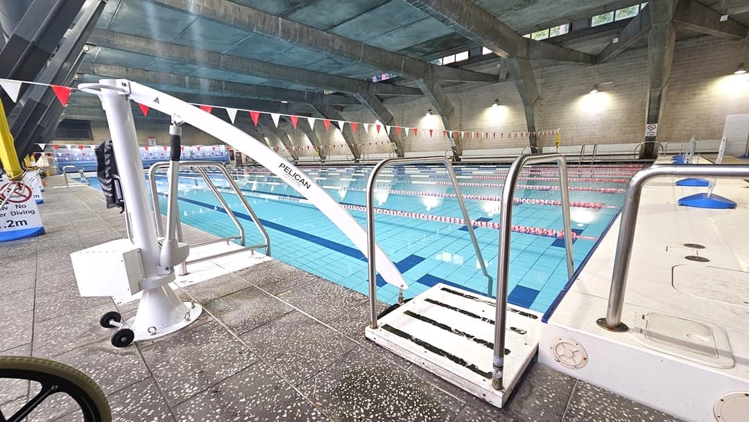 Indoor swimming pool with multiple lanes, separated by red and white lane dividers. A pool lift labelled "PELICAN" is on the left, mounted on a speckled tile floor. Metal railings are near the pool's edge. The ceiling is high with exposed beams and overhead lights. The background shows a wall with more lights and a no diving sign. The water is clear, reflecting the ceiling's structure.