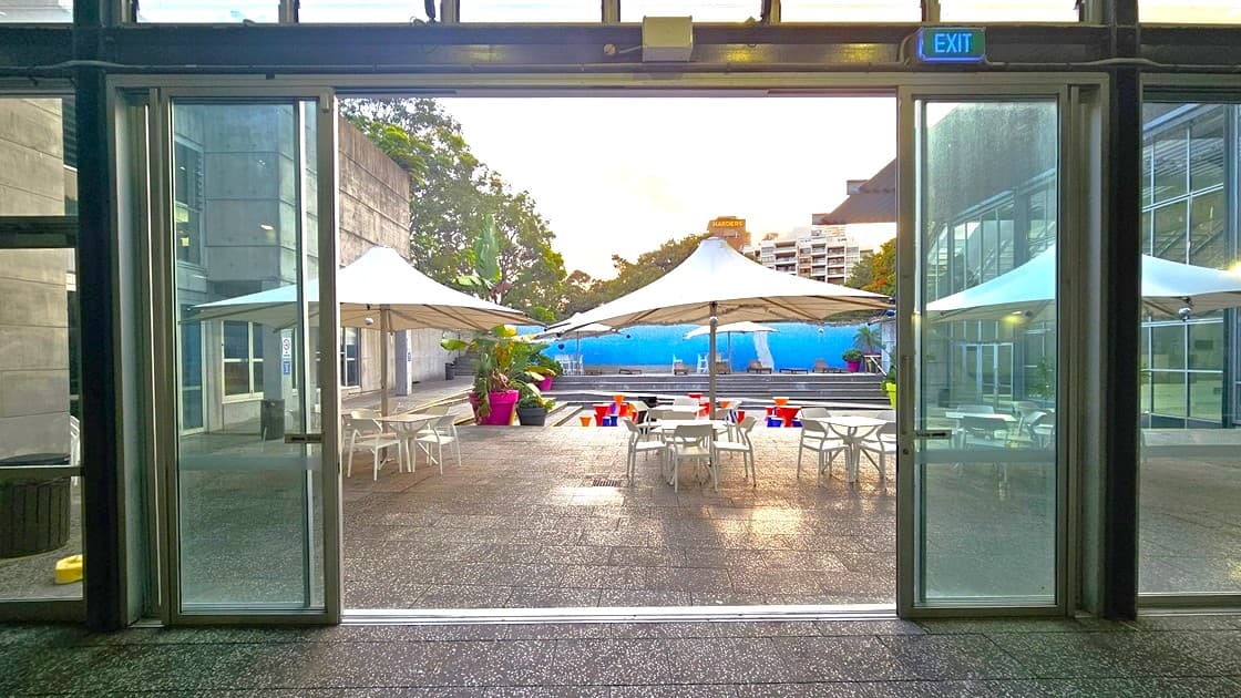 A view through large glass doors leading to an outdoor patio area. In the foreground, there are tables and chairs under large white umbrellas. The ground is paved with textured stone. In the background, there are colourful chairs and a blue wall, with trees and buildings visible beyond. The ceiling inside is not visible, but an "EXIT" sign is above the doors.