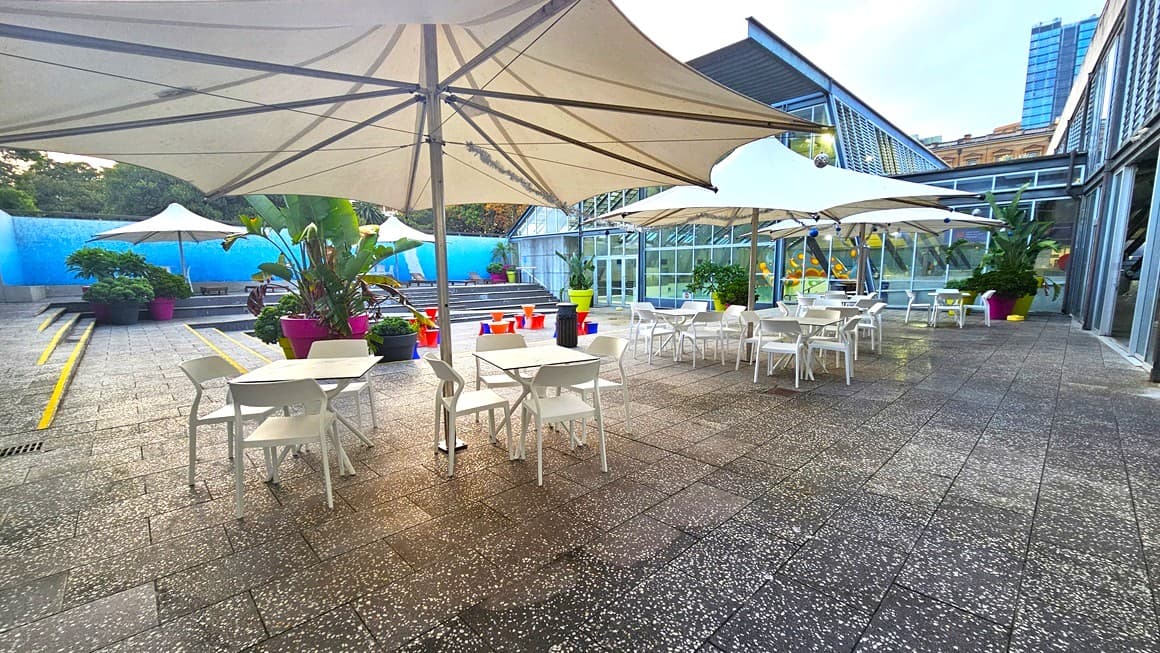 Outdoor seating area with white tables and chairs under large beige umbrellas. The ground is paved with speckled tiles. On the left, there are potted plants and a blue wall. In the background, steps lead up to more seating with colourful planters. On the right, a glass building with reflective windows and more potted plants. A tall building is visible in the distance.