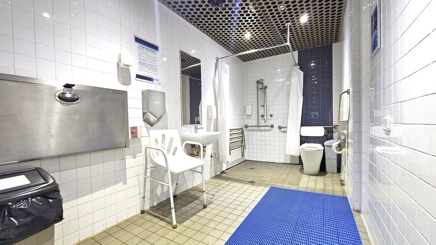 A spacious, accessible bathroom with white tiled walls and a perforated metal ceiling. On the left, there's a baby changing station and a waste bin. In the centre, a white chair is positioned next to a sink with a mirror above it. The right side features a toilet with grab bars and a shower area with a curtain and additional grab bars. The floor is tiled with a blue mat near the entrance.