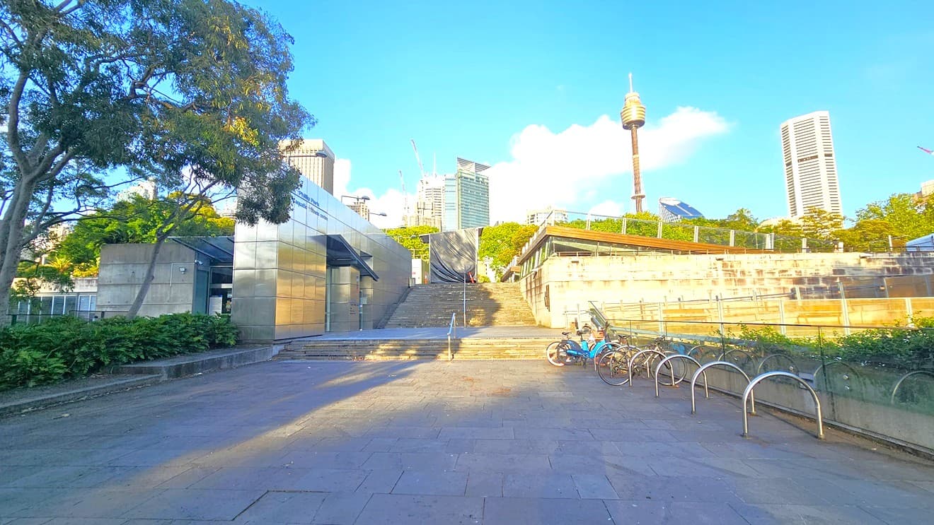 To the left is the facility building with silver walls. On the right, a bike rack holds a couple of bikes. In the middle, there are steps with a single handrail, leading to another set of steps for the upper level. Landscape ornamental plants and trees fill the plant box on the left. Tall trees and commercial buildings, including the Sydney Eye Tower, are visible.