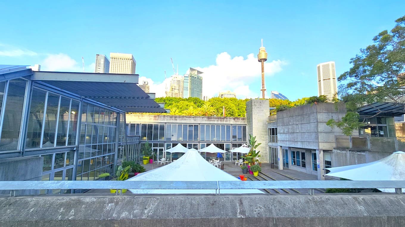 To the left is a building with floor-to-ceiling glass walls. In the middle is another building with glass walls, beneath umbrella shades, with arranged tables and chairs. Large plants in large pots are also visible. To the right is a concrete building with glass walls. The building's entrance features an extended glass roofing. Tall buildings and trees surround the area.