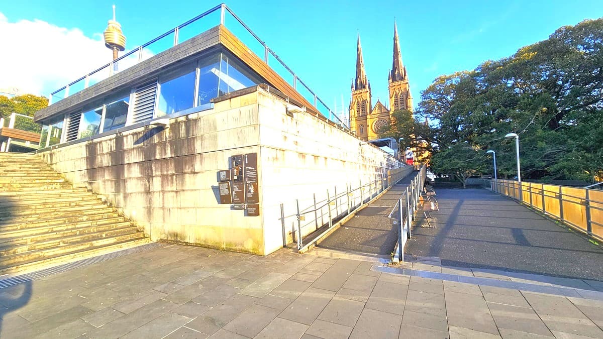 In the middle is the facility building with brick and glass walls featuring brown plates with historical data. On the right, there are two sets of elevating ramps with handrails. The church is visible on top of the ramps. On the left are concrete steps equipped with truncated domes. Tall trees and commercial buildings, including the Sydney Eye Tower, are visible.