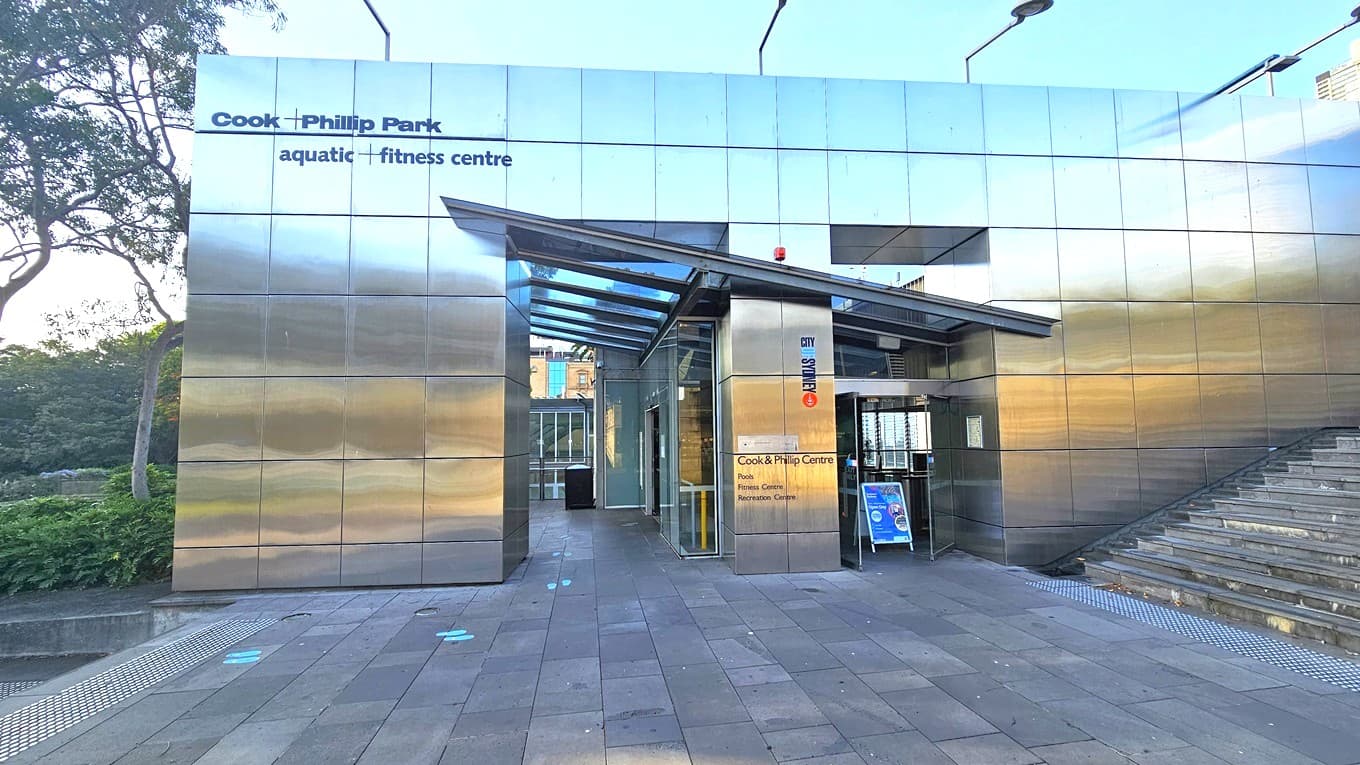 Entrance to the facility. In the middle is a building with silver walls, bearing the facility name on the upper left. Two entrances have an elevated glass roofing, with lights affixed to the top of the structure. Banners and posters are visible. To the left is an elevating stairs equipped with truncated domes. Trees and ornamental plants adorn the left side of the building. The floor has grey concrete tiles.
