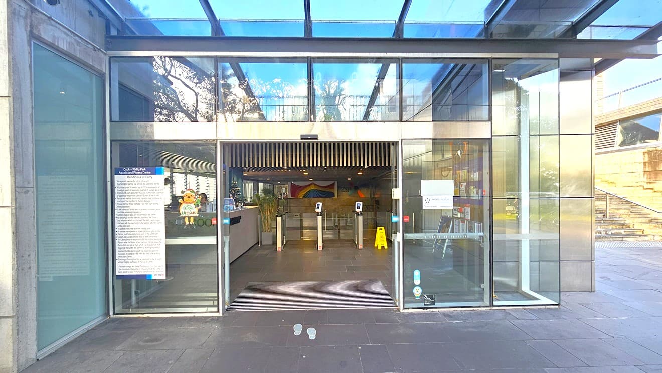 To the left is a retail display of swimwear, goggles, and swimming accessories. In the middle is the reception featuring the facility's name in front of the white and brown counter. Christmas decorations adorn the reception counter. To the right is a glass turnstile with a swiping machine. A large bunting is hung in the middle, near the timber ceiling. The ceiling features multiple looks of timber, grill, and linear designs. The space is well-lit with natural light from the glass ceiling above.