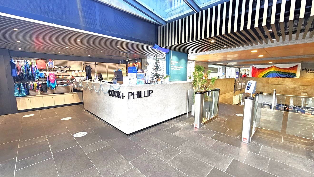 To the left is a retail display of swimwear, goggles, and swimming accessories. In the middle is the reception featuring the facility's name in front of the white and brown counter. Christmas decorations adorn the reception counter. To the right is a glass turnstile with a swiping machine. A large bunting is hung in the middle, near the timber ceiling. The ceiling features multiple looks of timber, grill, and linear designs. The space is well-lit with natural light from the glass ceiling above.