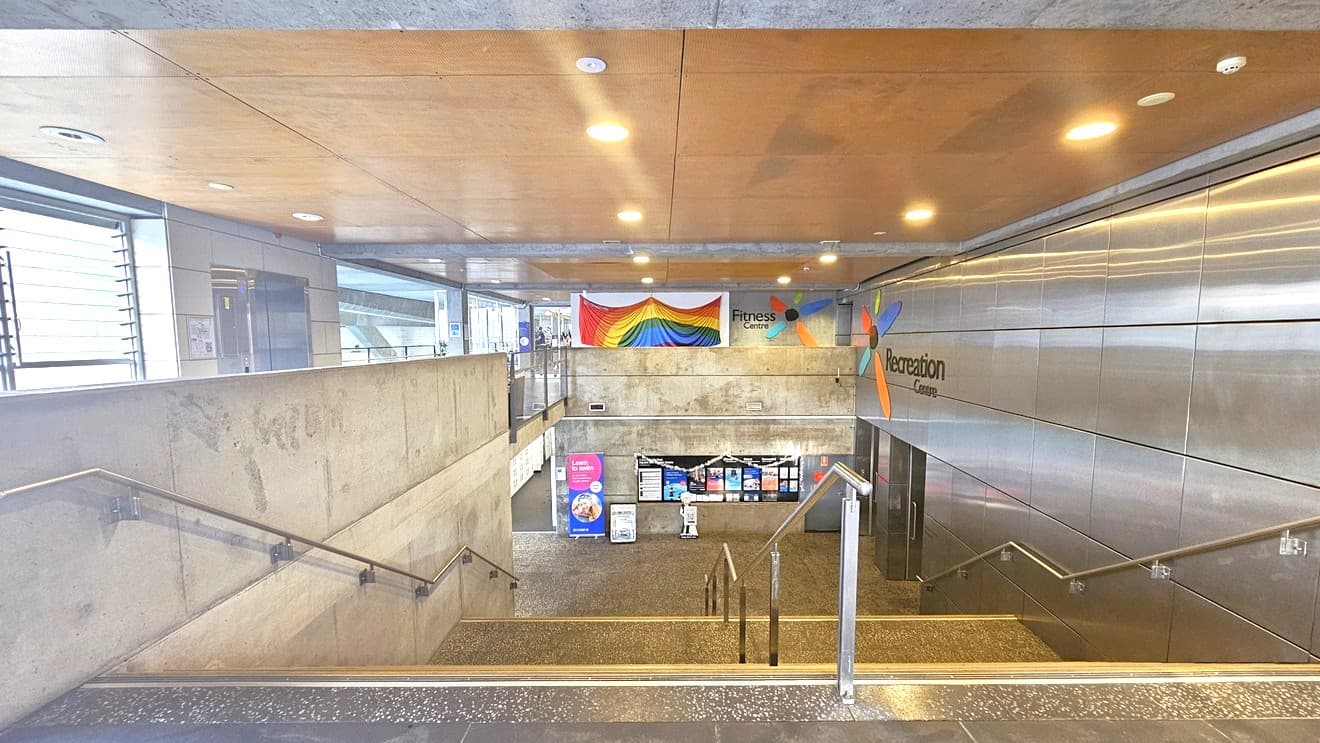 The stairs descend, leading to the facilities below. Both sides are equipped with handrails and an additional one in the middle. The steps have grey tiles with anti-slip aluminum alloy for safety. The ceiling has a timber design with LED lights. On the left is a corridor that leads to the gym. To the right is a wall with silver tiles bearing the facility's name. A large bunting flag is hung in the middle. Banners are displayed at the end of the stairs below.