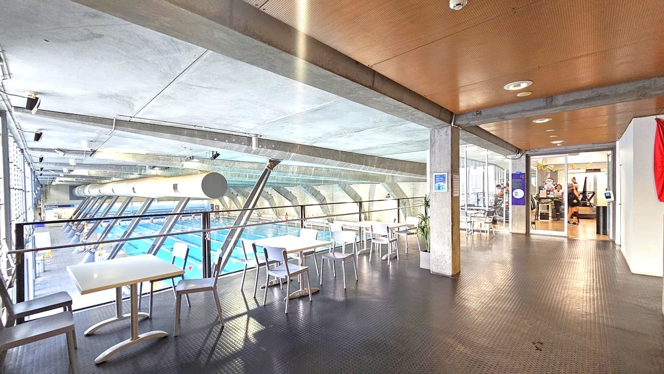 To the left, movable white tables and chairs are arranged. To the right is a glass door that leads to the gymnasium. A swiping machine and a banner are affixed to the wall. The floor features rubber tiles, and the timber ceiling has LED lights. Gym users are visible from inside the gym. To the left, the aquatic facility below is visible.