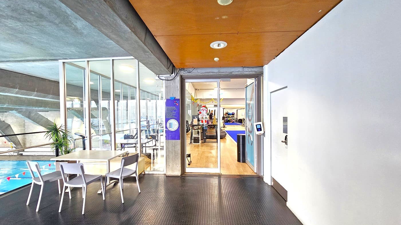 To the left is a set of movable plastic tables and chairs. The entrance to the gym is a manual inward-opening glass door equipped with a banner on the right side of the door. A swipe access point is affixed to the wall on the right. The floor transitions from black rubber tiles outside to timber. Treadmills are visible from the inside. The ceiling outside has a brown wooden design; on the left, the aquatic facility is visible below.