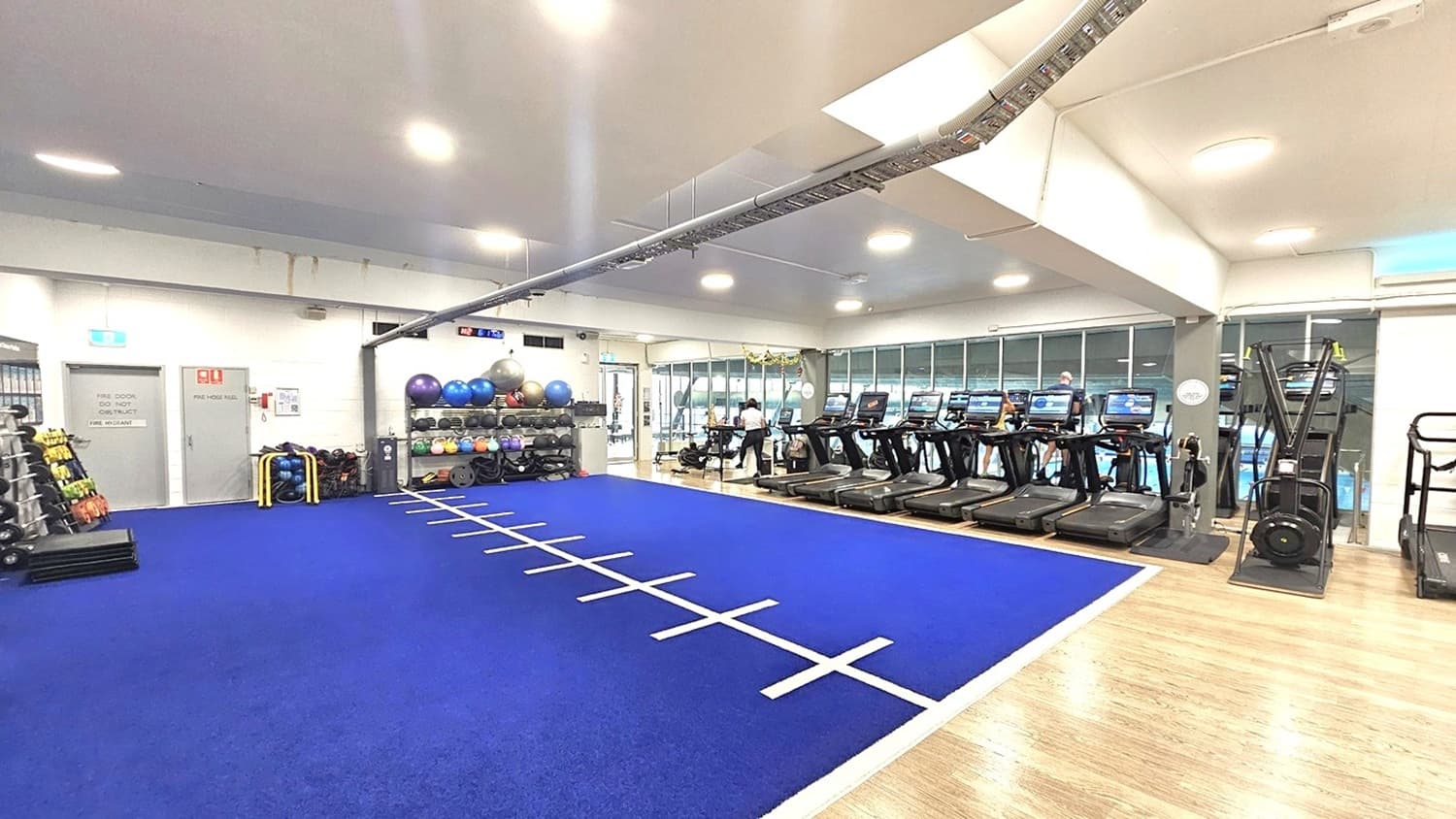 Inside the gymnasium, a blue soft flooring with a sprint track covers the left section. Weights, kettlebells, medicine balls, and stability balls are organized on the left. Two grey doors, along with a fire extinguisher, are visible. On the right, there are treadmills and a stairmill. The floor on the right changes to timber. The ceiling features a white design with LED lights and a metal structure affixed to the ceiling.
