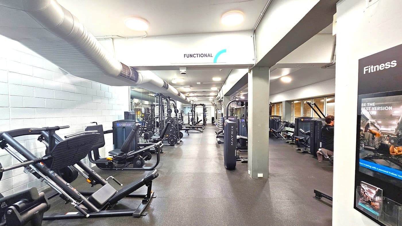 On the left, Smith Machines and Bench Press Machines are arranged. To the right, there are more strength machines. The floor is covered with grey rubber tiles, and the ceiling features an industrial design with exposed large beams and air conditioning vents. The left wall has a white brick design, while the right wall is adorned with floor-to-ceiling glass.
