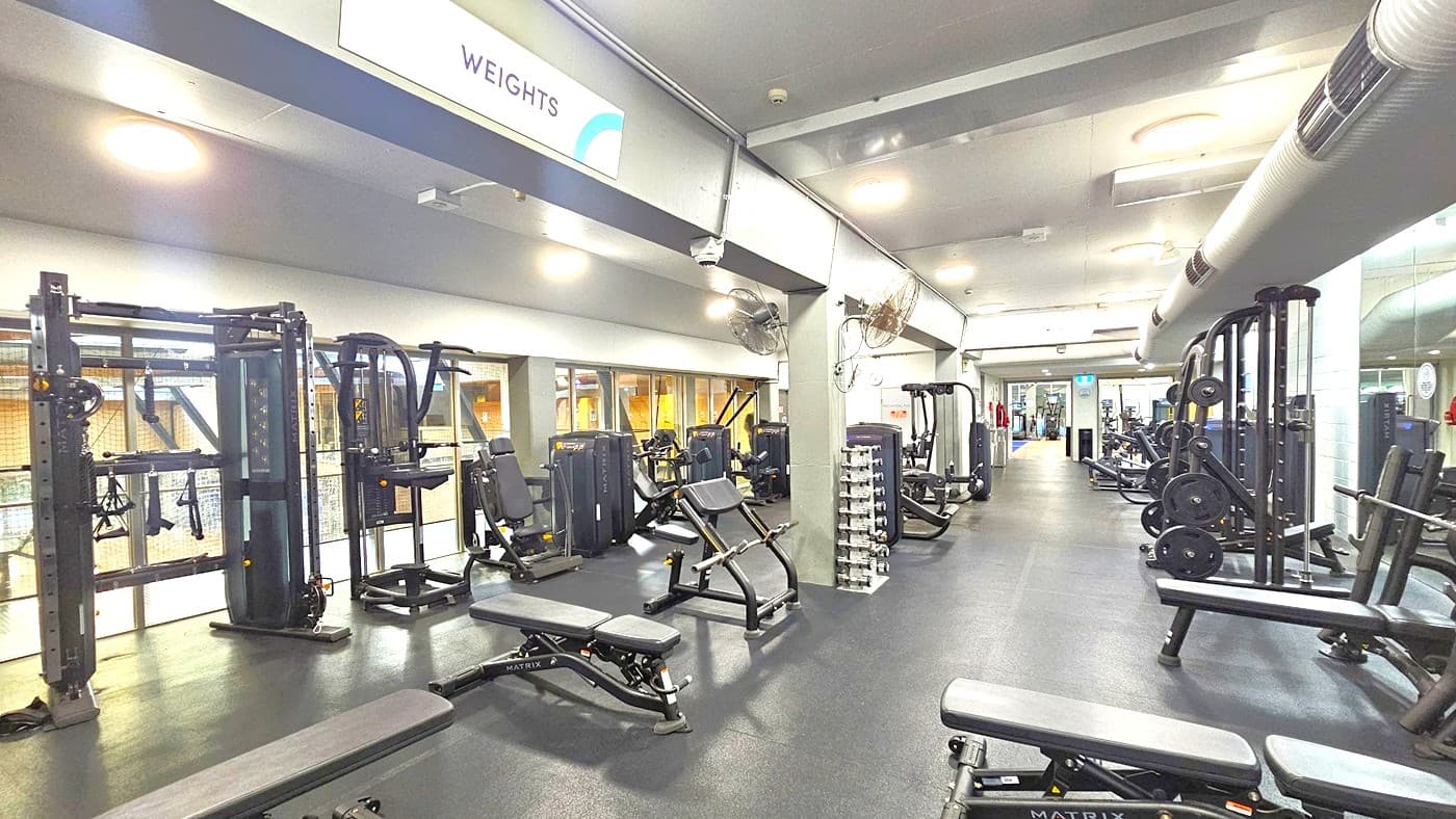 On the left, there are Smith machines, Bench Press, and Inclined Press Machines. In the middle, free weights are arranged. On the right, there are Lat Pulldown Machines and more Bench Press Machines. The floor is covered with black rubber tiles, and the ceiling is white with LED lights and large air conditioning vents. A ceiling fan is mounted on the middle wall. Glass walls surround the left.
