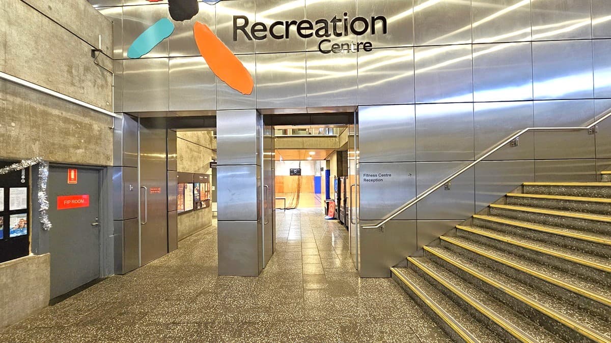 In the centre, two manual glass doors stand to the left of a silver wall, bearing the facility name and logo at the top. On the left, a grey door displays a red sign and garland decoration. To the right, a concrete staircase with handrails is present, featuring anti-slip aluminum alloy on each step for safety. The floor, including the steps, is adorned with a speckled design.