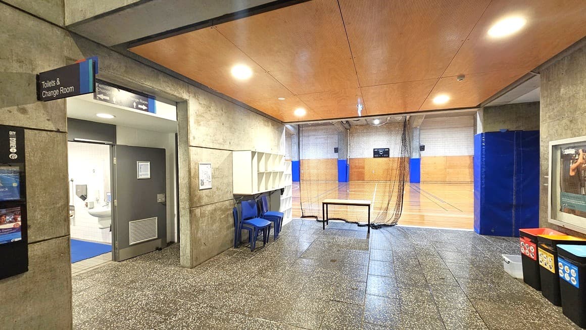On the left, an accessible Toilet and Change Room is accessed through a grey manual door. Signage and posters adorn the wall. A middle wall divides the bathroom from the corridor, where blue plastic chairs and white storage racks are placed. To the right, a large poster in a glass frame is displayed, accompanied by three black rubbish bins below. The wall features grey concrete, and the ceiling exhibits a wooden brown design with LED lighting. The court's timber floor is visible.