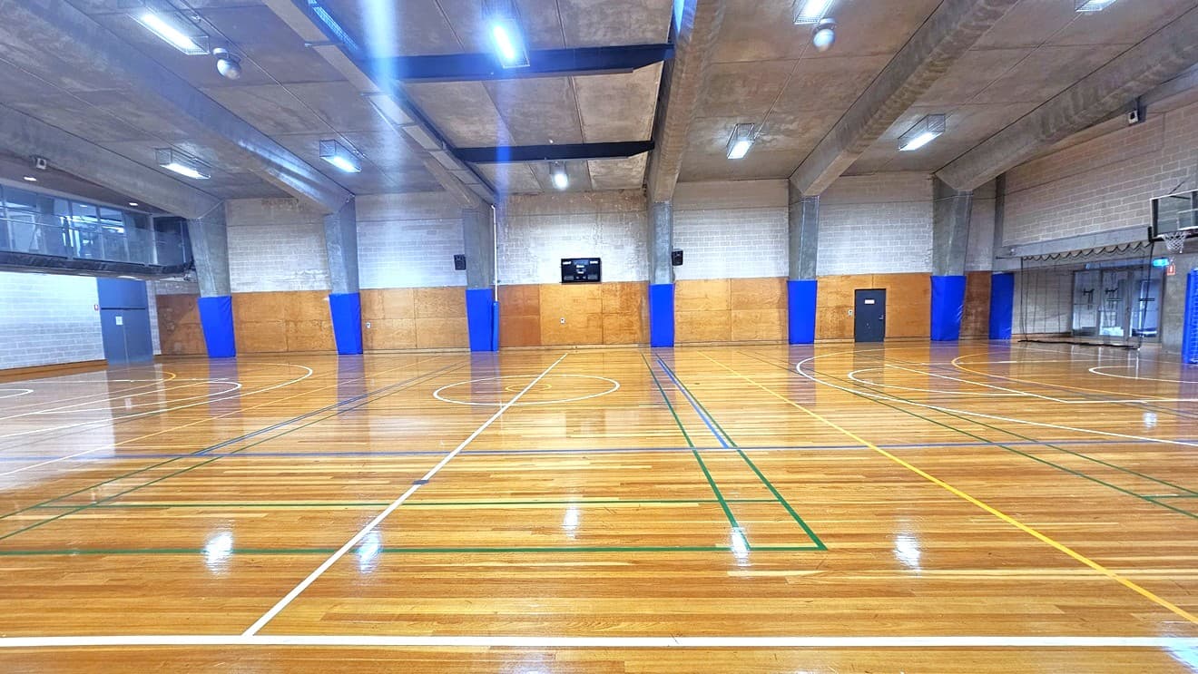 The court play area, constructed from timber, features markings for various ball games. A vantage point for spectators is situated on the upper left, enclosed by glass. The walls are built with bricks and adorned with blue accents. The ceiling has an industrial appearance, showcasing large concrete beams and fluorescent lights.