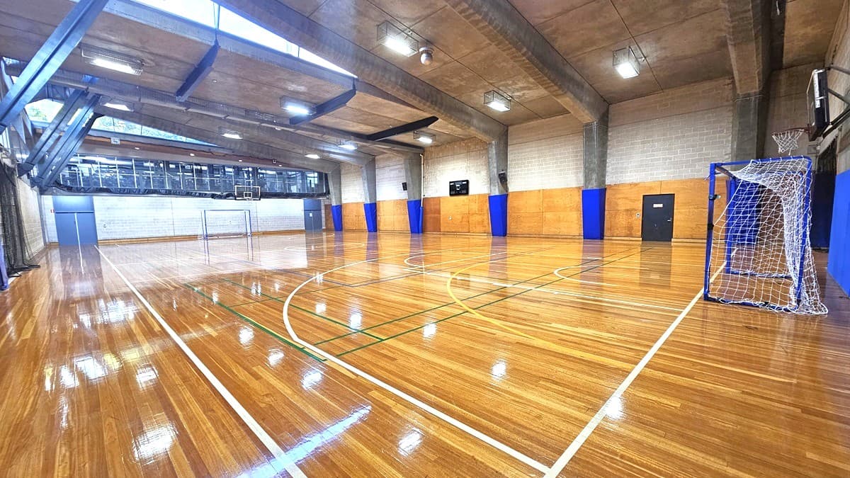 The court play area, crafted from timber, displays markings for various ball games. A spectator vantage point, enclosed by glass, is positioned on the upper left. The walls are constructed with bricks and decorated with blue accents. The ceiling adopts an industrial aesthetic, featuring large concrete beams and fluorescent lights. A goal is situated on the right beneath the basketball board. Black nets are suspended from the ceiling in the middle, creating a separation between the two courts.