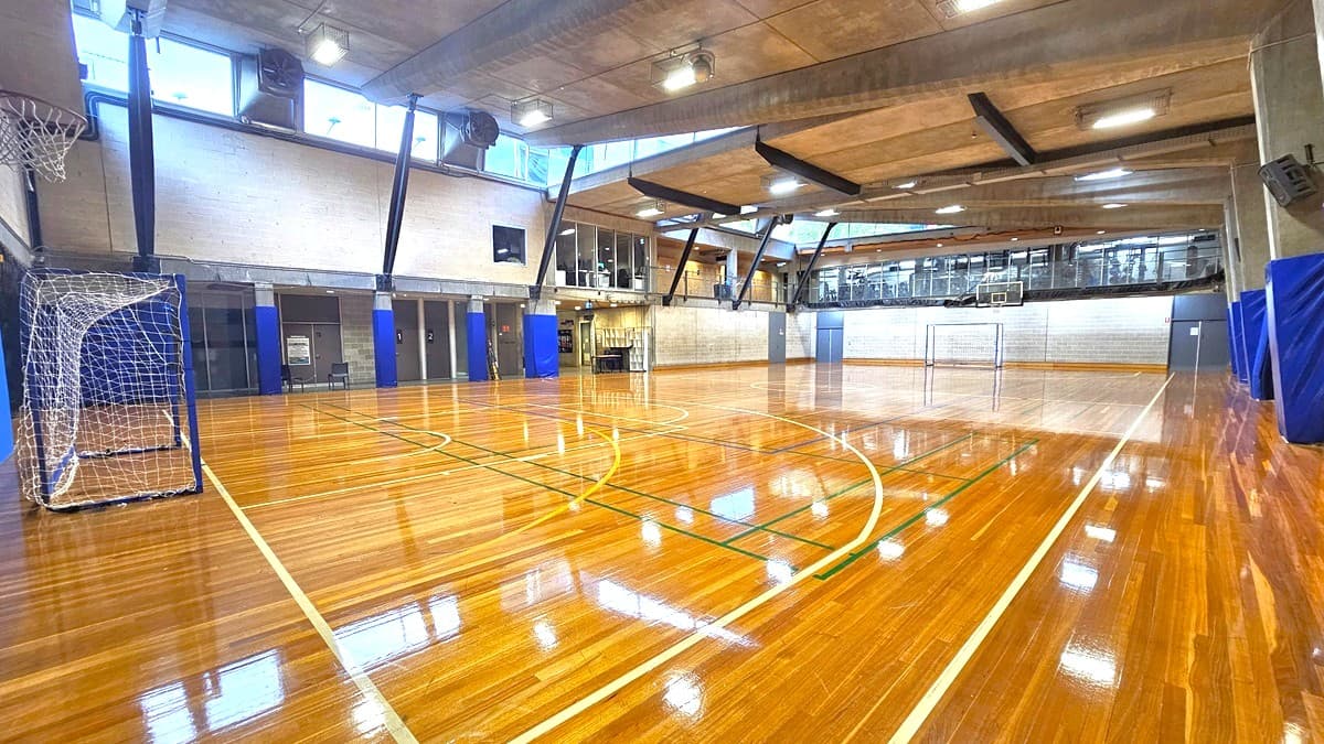 The court play area, crafted from timber, displays markings for various ball games. A spectator vantage point, enclosed by glass, is positioned on the upper right. The walls are constructed with bricks and decorated with blue accents. The ceiling adopts an industrial aesthetic, featuring large concrete beams and fluorescent lights. A goal is situated on the left beneath the basketball board. Black nets are suspended from the ceiling in the middle, creating a separation between the two courts.