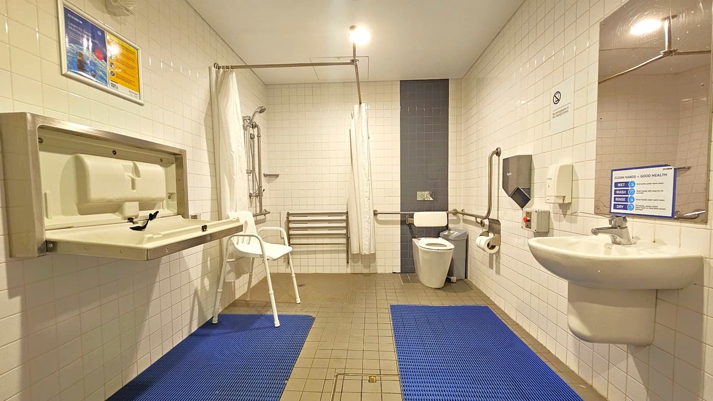 The bathroom includes a change table and a white plastic chair on the left. On the same side, a shower with hand grabs and a curtain is fixed. The right side features a toilet bowl with hand grabs and a rubbish bin. A washbasin, mirror, soap and tissue dispenser, and an emergency button are mounted on the right wall. Blue non-slippery mats cover the floor. Safety banners and reminders are affixed to the walls, which are clad in white tiles. The ceiling is uniformly white.