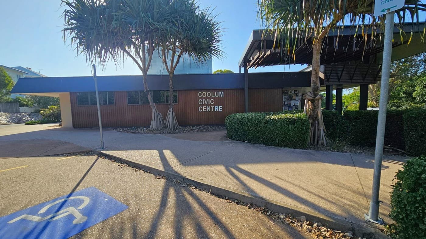 A street scene with a "No Through Road" sign on the left, surrounded by lush greenery and a large tree. The Coolum Civic Centre is in the centre, featuring a modern design with a slanted roof. To the right, more trees and a sign for Jack Morgan Park are visible. The pavement is a mix of concrete and asphalt, with shadows cast by the trees. The sun is partially visible, creating a bright spot in the sky.