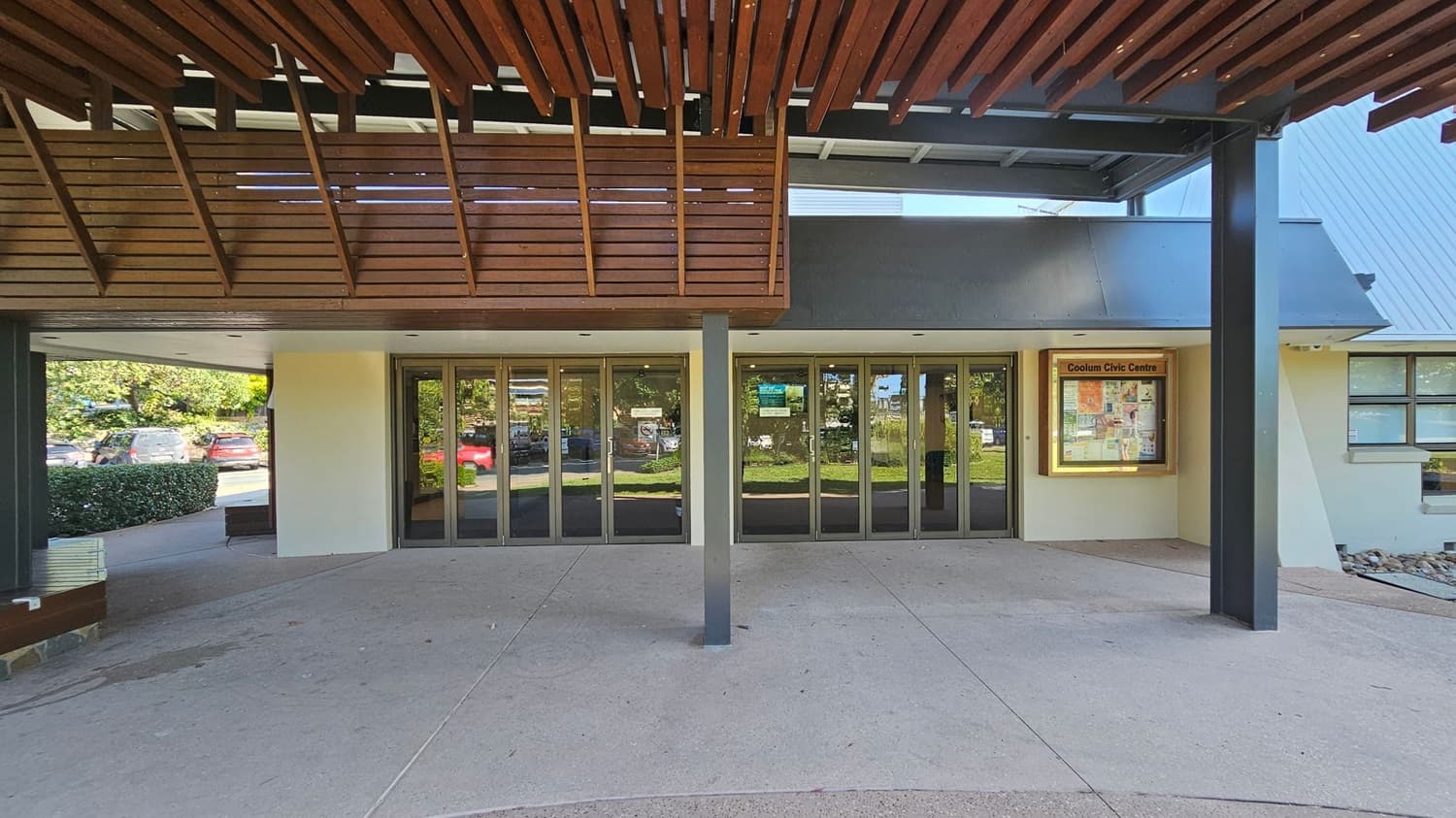 A building entrance with large glass doors in the centre, reflecting the greenery outside. Above the doors, a wooden slatted awning provides shade. To the right, a notice board displays information for the Coolum Civic Centre. The floor is a light-coloured concrete, and there are parked cars and bushes visible to the left through the glass. The ceiling features wooden slats matching the awning.