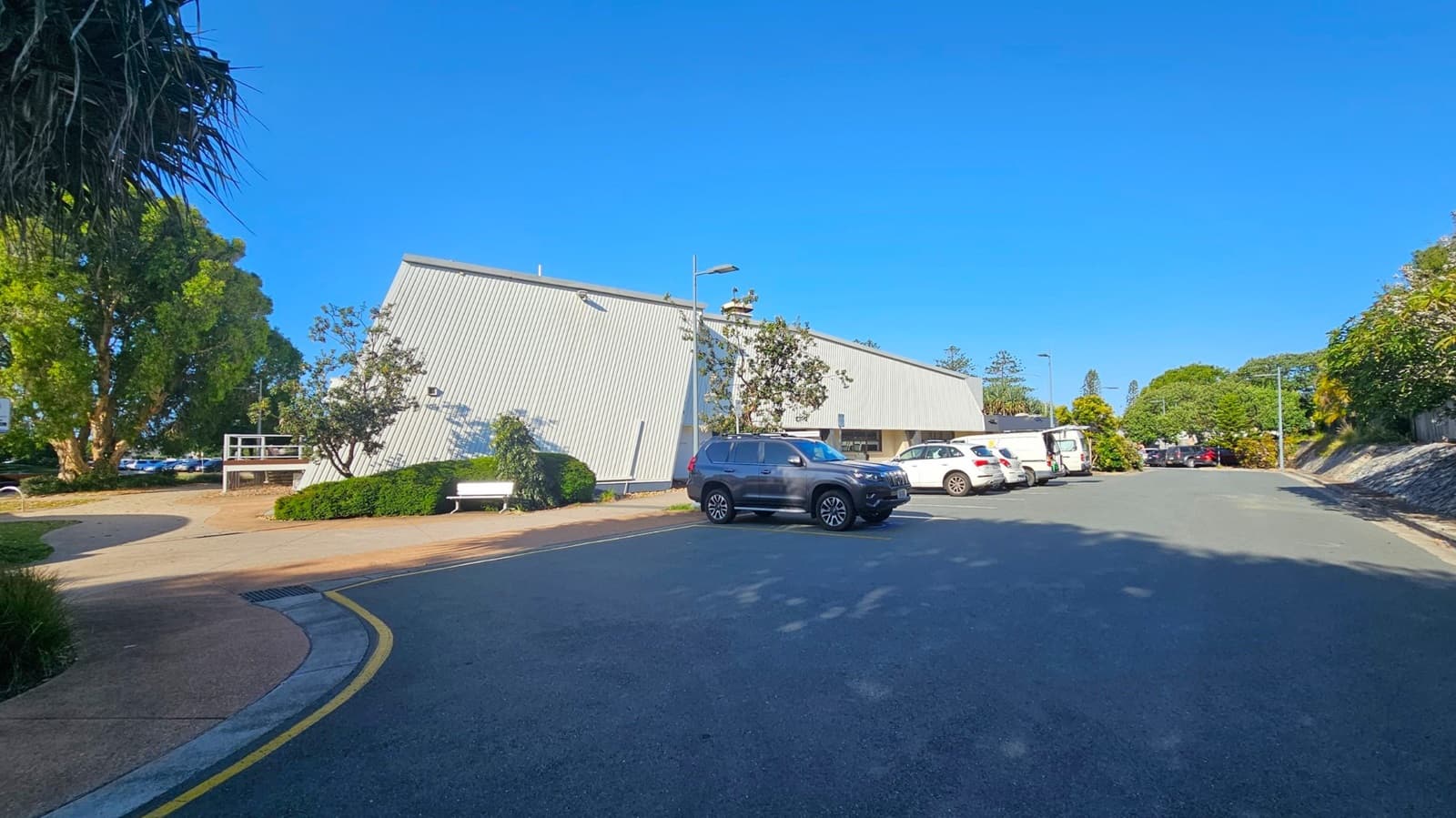 A large, modern building with a slanted roof is on the left, surrounded by trees and shrubs. A white bench is near the entrance. Several cars, including a black SUV, are parked in front of the building. The road curves slightly, with a yellow line marking the edge. More trees and greenery are visible in the background, lining the road.