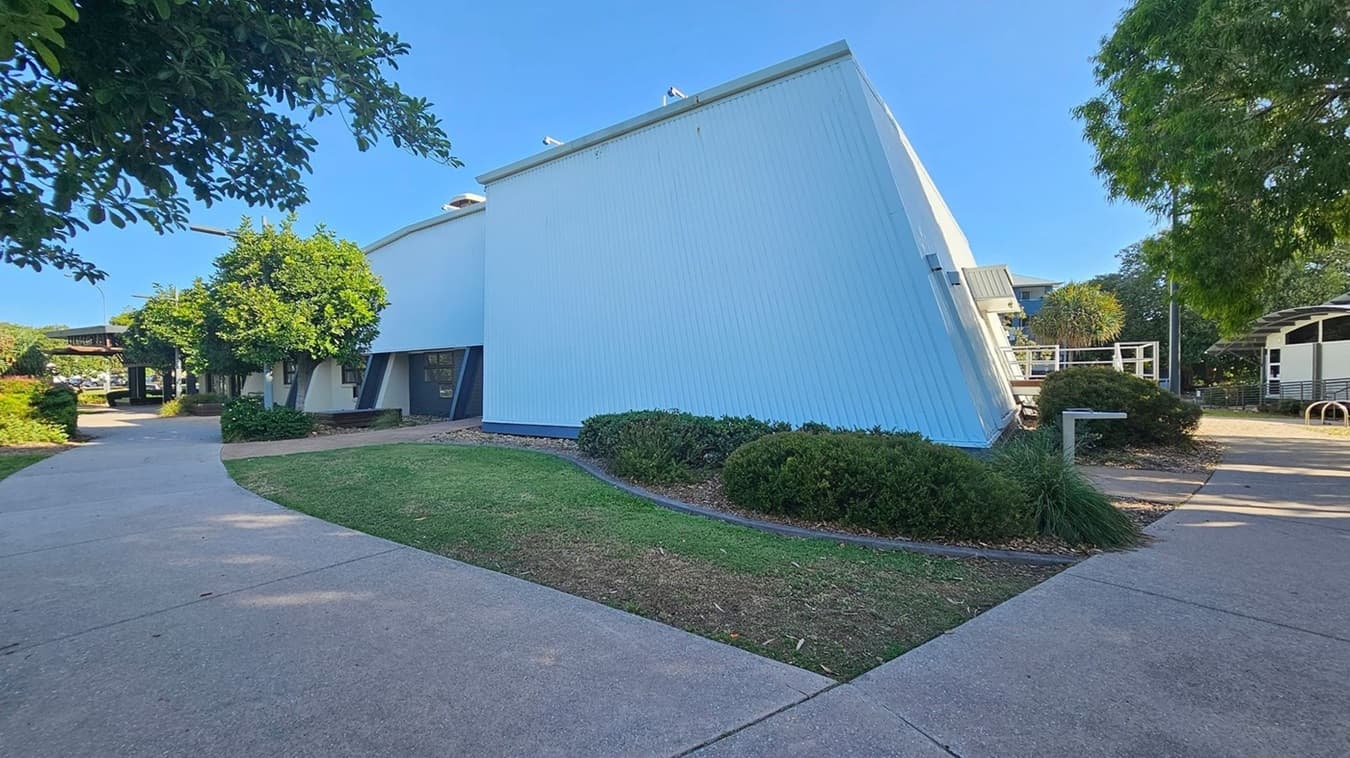 A modern building with a slanted white facade is on the right, surrounded by neatly trimmed bushes and a small lawn. A concrete path curves around the building, leading to a shaded area with more trees and foliage on the left. In the background, there are additional structures and a bicycle rack. The scene is well-lit, suggesting a sunny day.