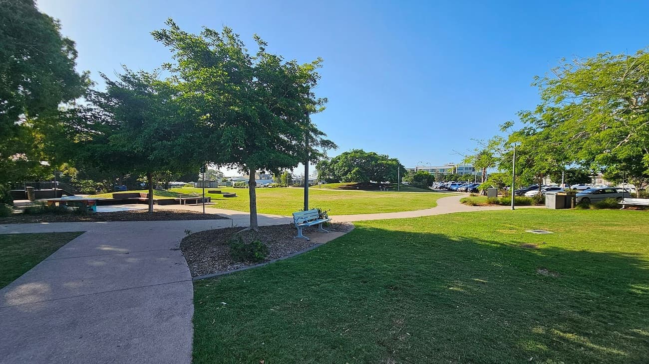 A park with a paved path curving through the centre. On the left, there are trees providing shade and a few benches. In the middle, a grassy area is surrounded by more trees. On the right, a row of parked cars is visible beyond the grass, with additional benches and trees lining the path. The sky is clear and blue.