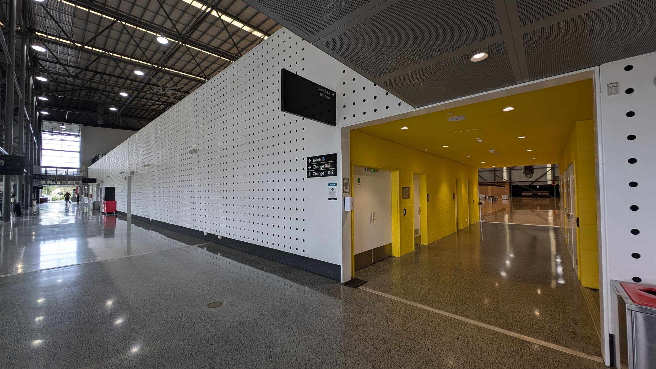 A spacious indoor area with a high ceiling and large windows on the left side. The floor is polished and reflective. On the right, there is a white wall with a pattern of black dots and a yellow entrance leading to a corridor with doors. Overhead lights illuminate the space. A sign near the entrance indicates directions to toilets and changing rooms. In the background, there are more windows and a few people walking.