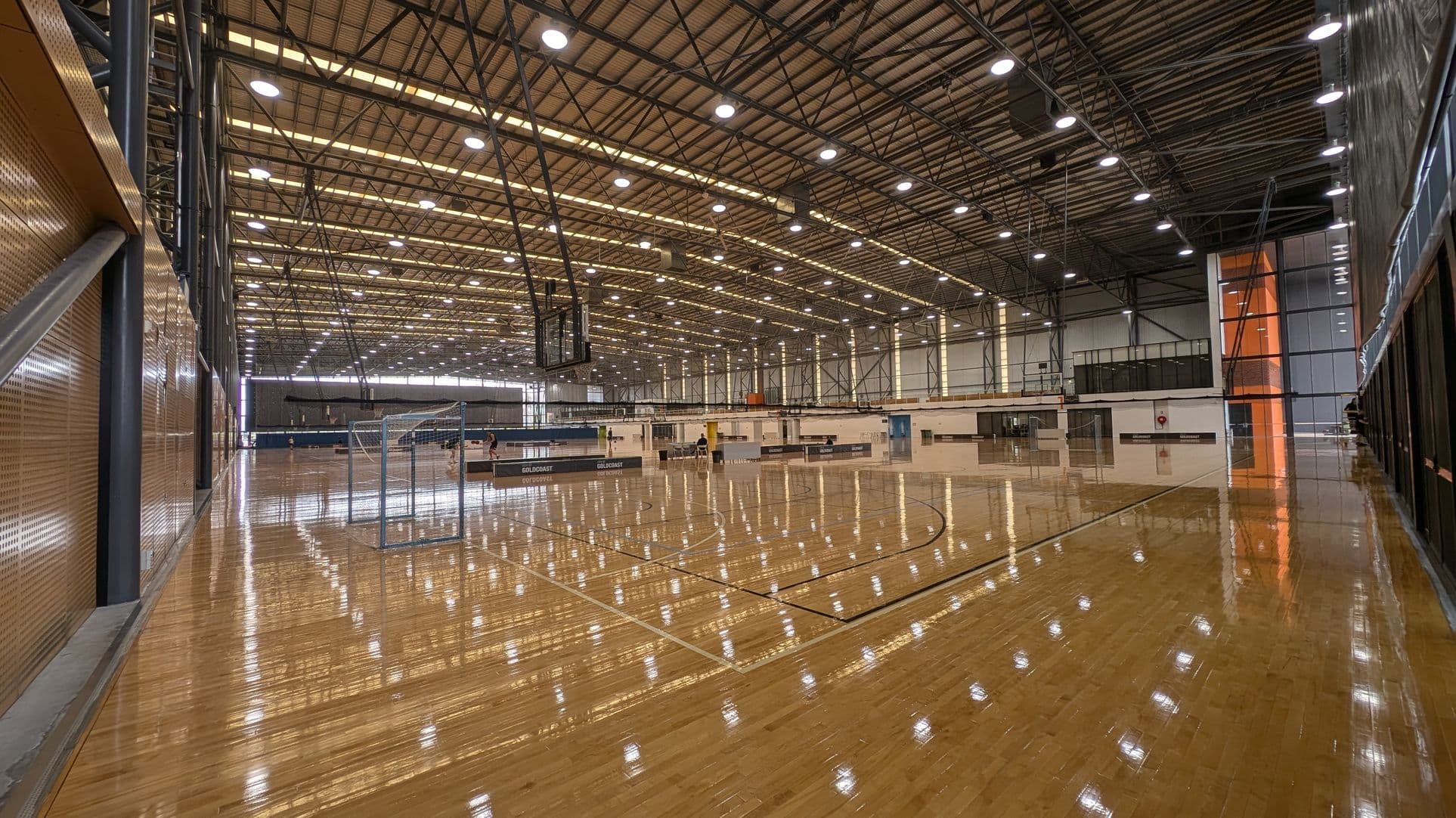 A large indoor sports hall with a polished wooden floor. On the left, there is a wall with metal beams and perforated panels. In the middle, there are multiple basketball courts with hoops and backboards. The ceiling is high with numerous lights and metal trusses. On the right, there are large windows and doors, with some areas leading to other sections of the building. The background shows more sports equipment and seating areas.