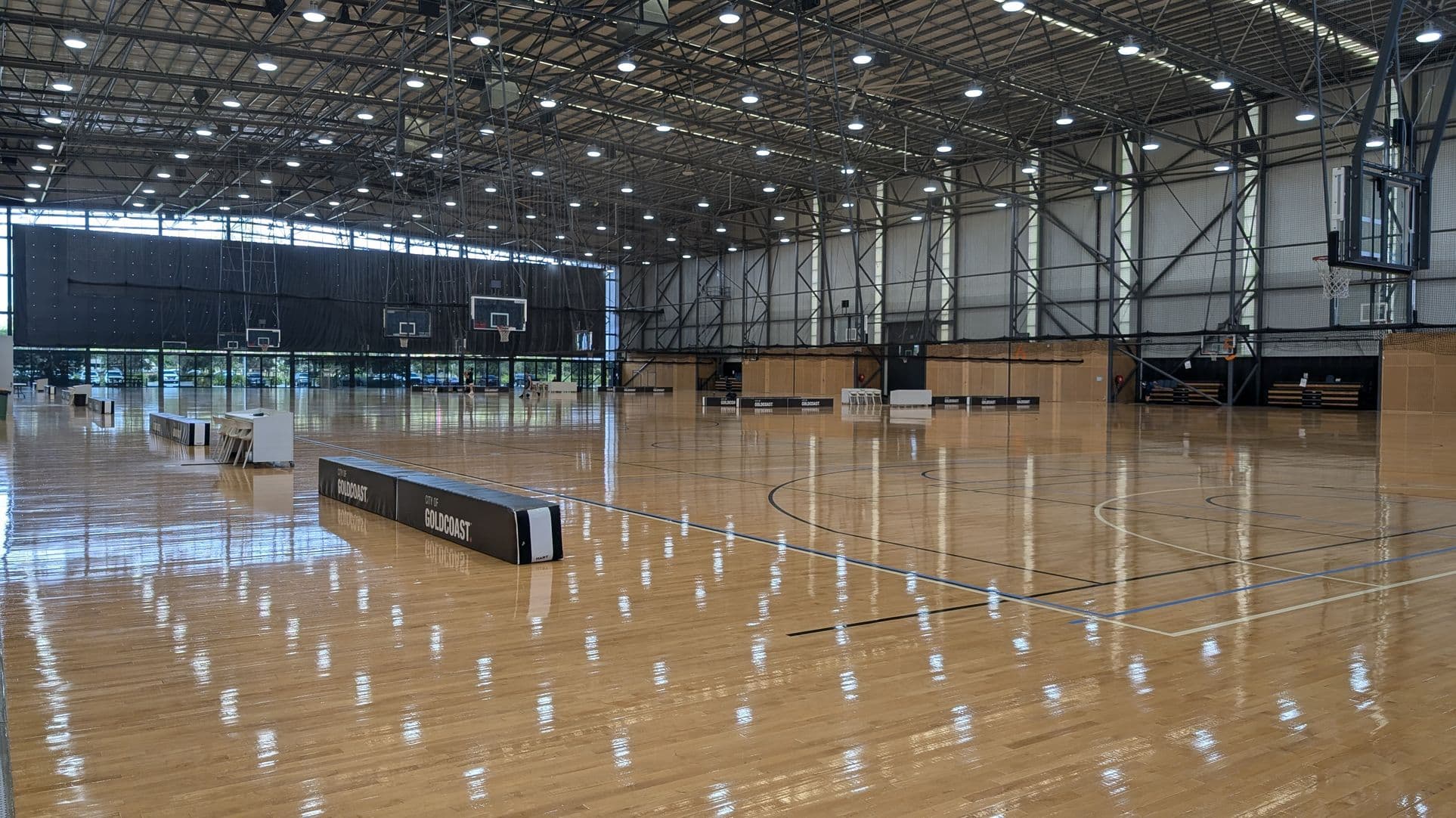 A large indoor basketball court with a polished wooden floor. Multiple basketball hoops are visible on the left and right sides. The ceiling is high with a metal framework and numerous lights. The background features large windows allowing natural light to enter. In the foreground, there are barriers with "CITY OF GOLD COAST" written on them. The walls are lined with light-coloured panels and some seating areas.