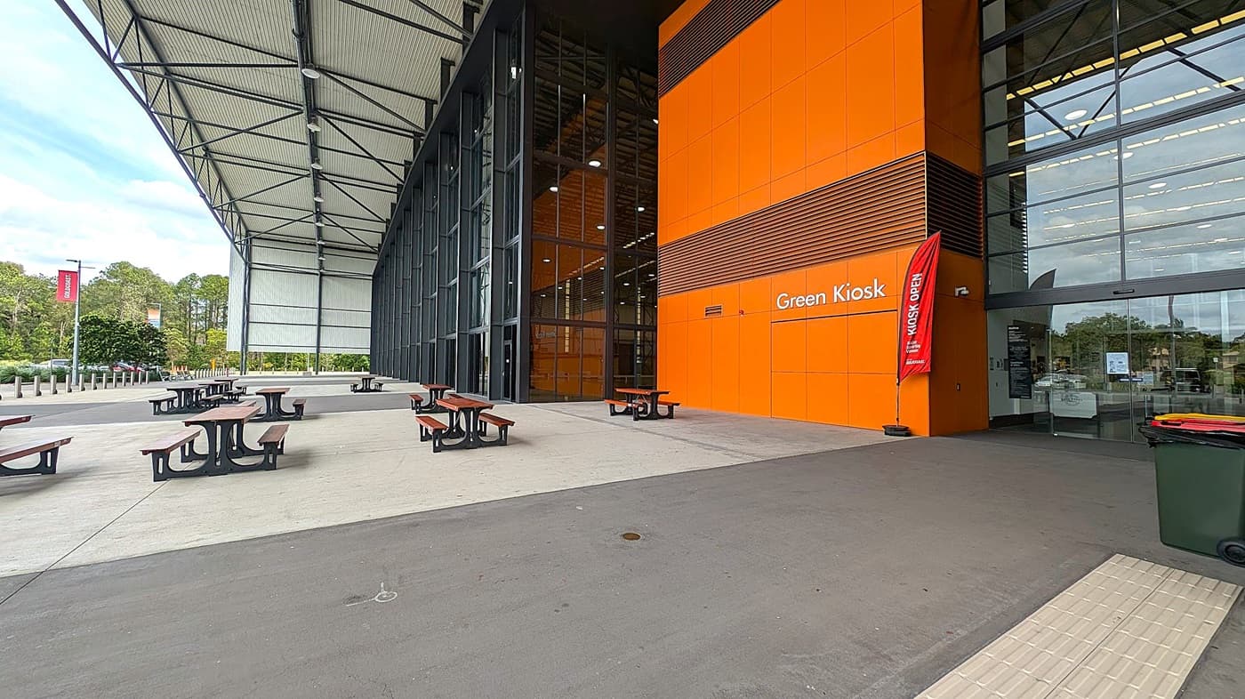 A large, modern building with an orange facade on the right side, labelled "Green Kiosk." A red flag next to the entrance reads "Kiosk Open." The entrance has large glass doors and windows. In front of the building, there are several wooden picnic tables with metal frames on a concrete surface. The left side shows a covered area with a high, metal roof supported by beams. In the background, there are trees and a few parked cars. The floor is concrete, and the ceiling is metal.