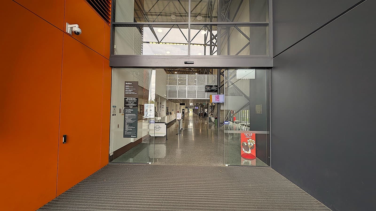 Entrance to a modern building with glass doors. On the left, there is an orange wall with a security camera mounted near the top. On the right, a dark grey wall. Inside, a long corridor with a shiny floor, a reception desk on the left, and a staircase on the right. Various signs and notices are posted near the entrance, and a digital screen is mounted on the right wall. The ceiling has exposed beams and lights.