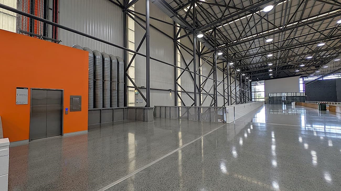A spacious industrial interior with a polished concrete floor and a high, metal truss ceiling. On the left, there is an orange wall with a metal lift door and a sign next to it. Adjacent to the wall are several large, grey ventilation ducts. The right side features a long, open area with metal support beams and large windows at the far end, allowing natural light to enter. The background includes a large, open space with additional windows and a few scattered objects.