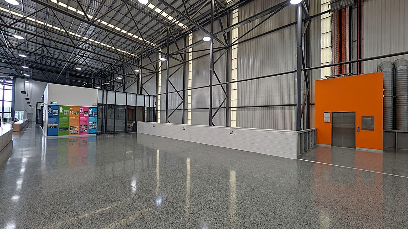 A spacious industrial interior with a polished concrete floor and a high, metal truss ceiling. On the left, there is a white structure with colourful posters displaying years and images. Adjacent to it is a long counter with hanging lights above. On the right, there is an orange wall with a metal door, possibly an elevator, and exposed pipes running vertically. The background features large windows and metal wall panels with vertical light strips.