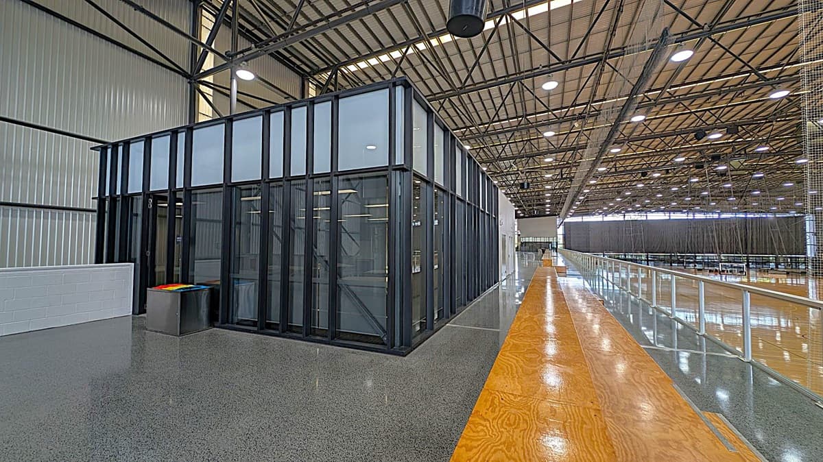 A large indoor sports facility with a high, metal truss ceiling and bright overhead lighting. In the foreground, there is a polished grey floor and a small area with wooden flooring. On the left, there is a black-framed glass structure with frosted upper panels and a set of colourful bins. To the right, a long walkway with a glass railing extends into the background, overlooking multiple sports courts. The walls are made of corrugated metal.