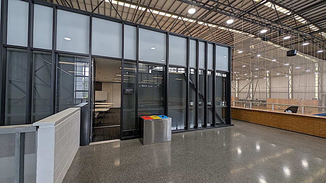 A modern indoor space with a polished grey floor and a high ceiling with exposed beams and lights. On the left, there is a glass-walled room with a door, partially open, revealing a meeting room with tables and chairs. In front of the glass wall, there is a recycling bin with separate compartments for different types of waste. On the right, there is a wooden railing overlooking a large open area with industrial-style metal framework and additional lighting.