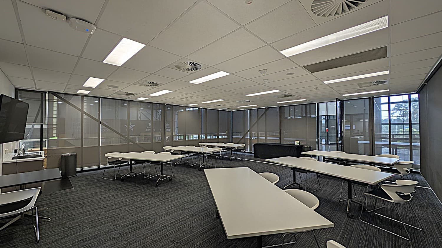 A modern classroom with grey carpeted flooring and a white tiled ceiling featuring recessed lighting and circular air vents. The room has several white tables and chairs arranged in clusters. On the left, there is a wall-mounted television and a small desk with a chair. The right side has large windows with roller blinds, allowing natural light to enter. The background shows a black cabinet and more tables and chairs near the windows.