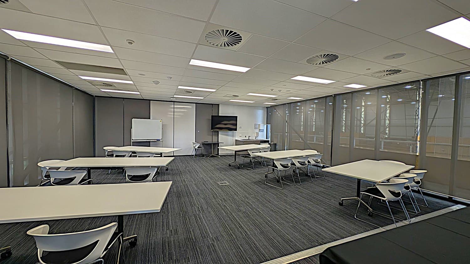 A modern classroom with grey walls and a dark grey carpeted floor. The ceiling is white with recessed lighting and circular air vents. Several rectangular tables with white tops and black legs are arranged around the room, each accompanied by white chairs. At the front, there is a whiteboard and a large flat-screen TV mounted on the wall. The right side of the room features large windows with grey blinds.