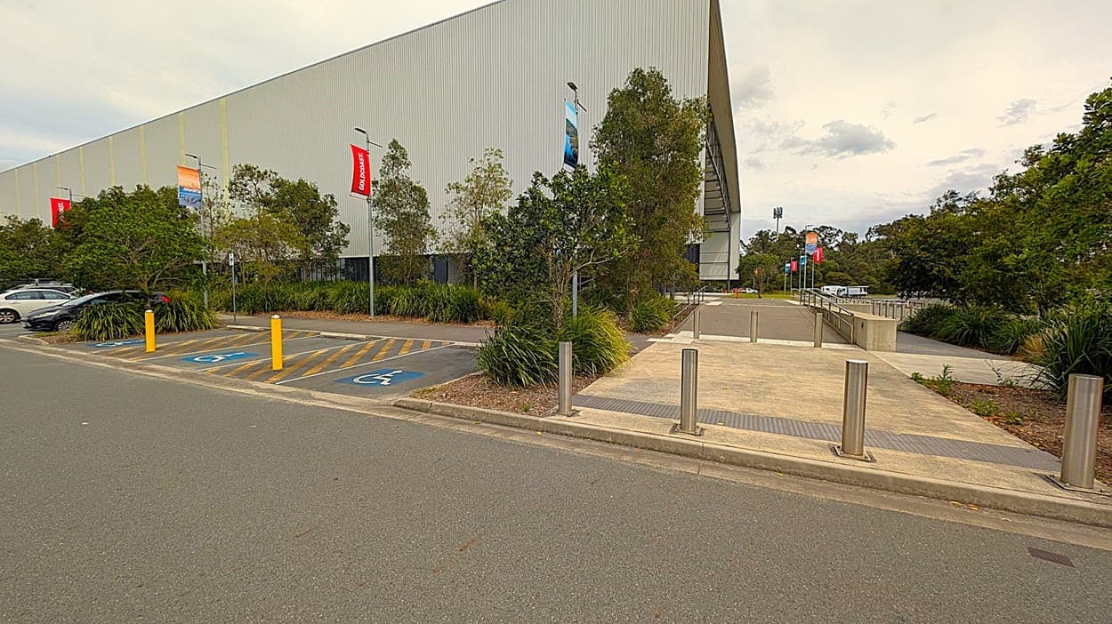 A large grey building with a corrugated metal exterior is on the left. In front of the building, there are several trees and bushes. There are three parking spaces marked for disabled access, with yellow-striped lines and blue wheelchair symbols. Yellow bollards are positioned at the front of the parking spaces. On the right, there is a concrete pathway with metal bollards and a ramp leading to the building. The background features more trees and a few parked cars.