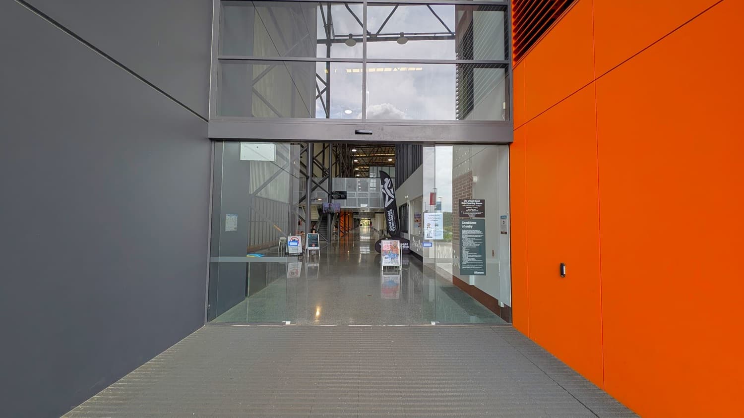 Entrance to a modern building with large glass doors. The left wall is dark grey, while the right wall is bright orange. Inside, the floor is polished and reflective. Several signs and posters are visible, including a black flag with white text and a green sign listing conditions of entry. The ceiling has exposed beams and lighting fixtures. The background shows a long corridor with additional signage and a glimpse of greenery outside.