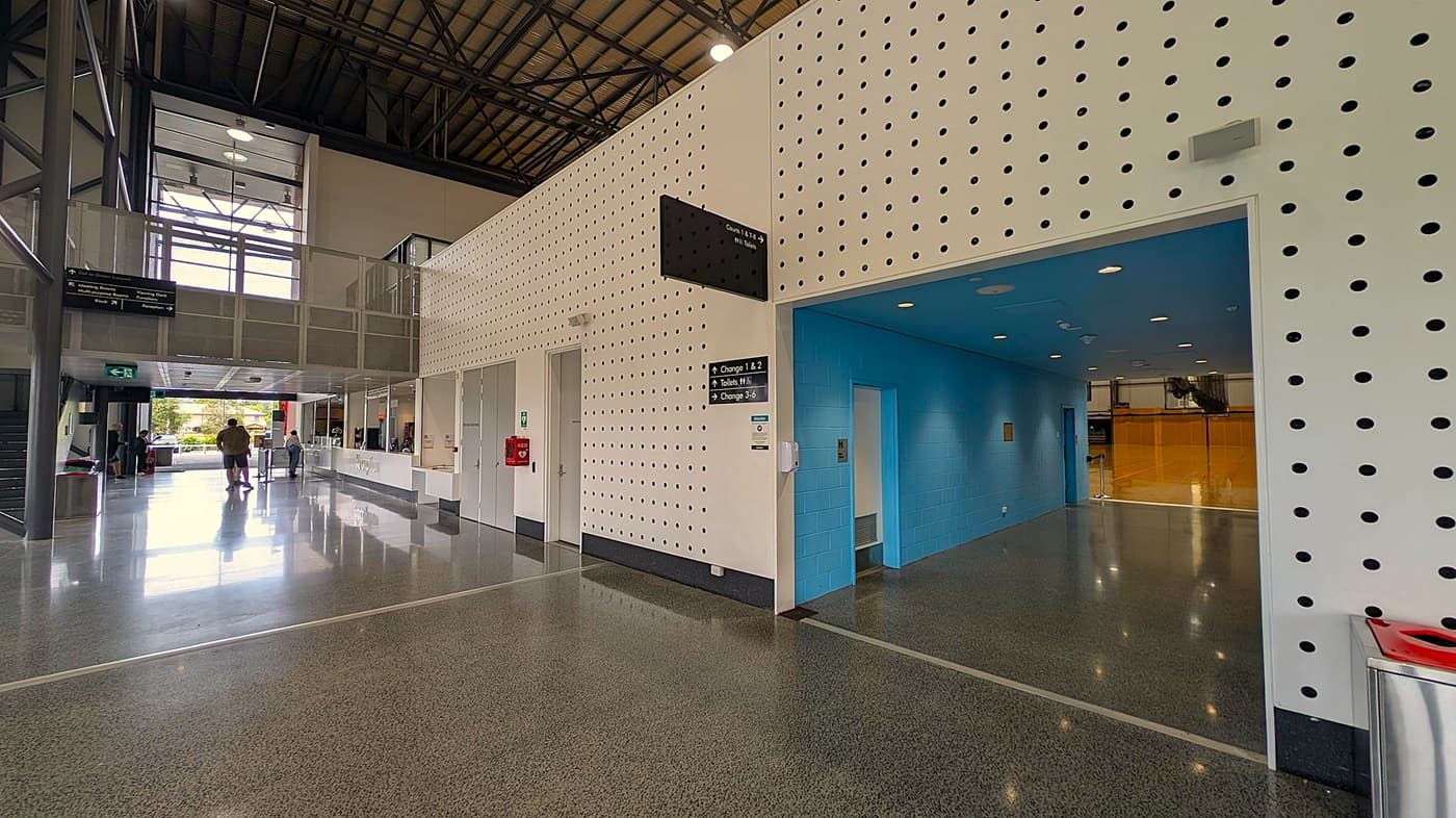 A spacious reception area with a white desk on the left, featuring the word "Reception" embossed on it. Behind the desk are several computer monitors, a wall clock, and a large screen displaying an image. The floor is polished grey with green speckles, and the ceiling is grey with recessed lighting. On the right, there is a long corridor with white perforated walls, a fire extinguisher, and an emergency exit sign. The corridor extends into a well-lit area with large windows.
