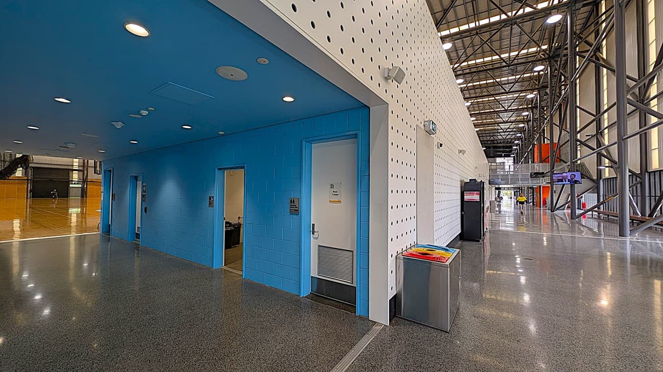 A spacious indoor area with a polished grey floor and a high ceiling with exposed beams and lights. On the left, there are blue-walled rooms with white doors, one labelled "Accessible Toilet." In the middle, a white perforated wall extends along the corridor. On the right, a long hallway with metal beams and large windows. A colourful recycling bin is near the white wall, and a person in a yellow shirt is walking in the distance.